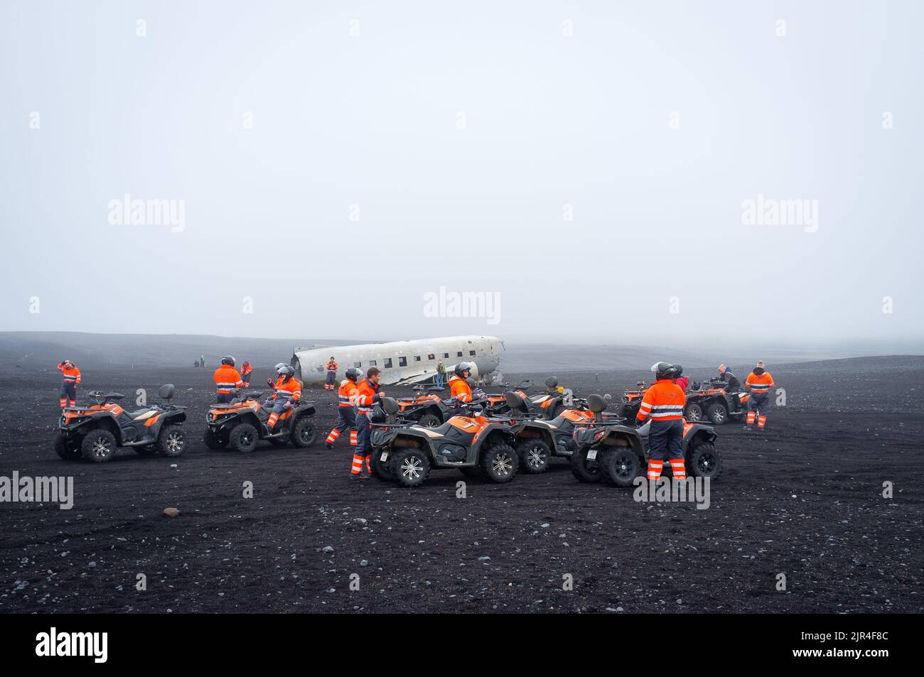Touristes dans des combinaisons d'orange sur des quads près de l'épave abandonnée de DC plane sur Solheimasandur en Islande un jour brumeux Banque D'Images Touristes dans des combinaisons d'orange sur des quads près de l'épave abandonnée de DC plane sur Solheimasandur en Islande un jour brumeux Banque D'Images