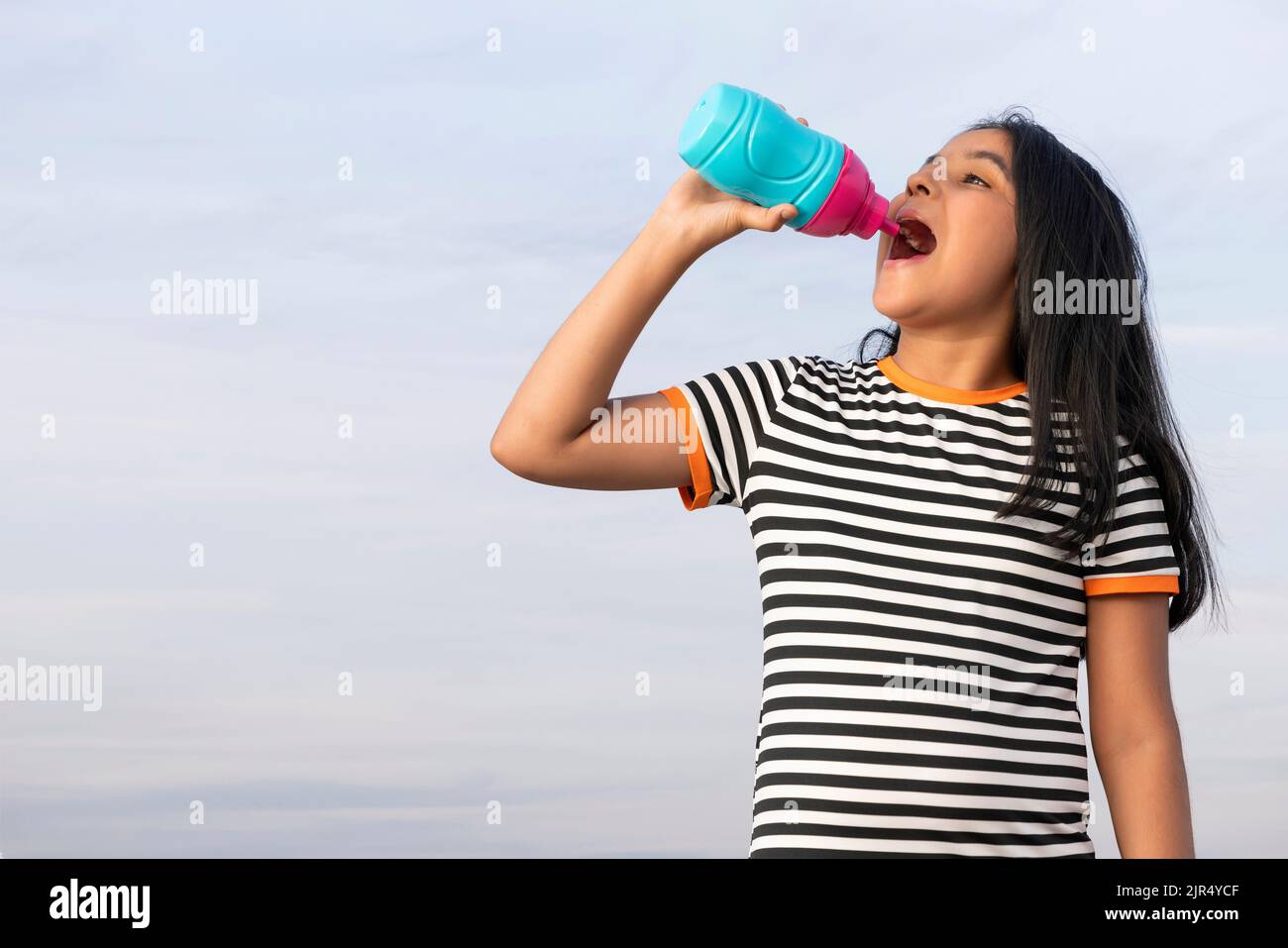 Une fille assoiffée de boire une bouteille d'eau tout en se tenant contre le ciel bleu Banque D'Images