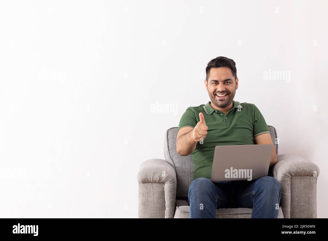 Jeune homme joyeux montrant un geste du pouce assis sur un canapé avec un ordinateur portable sur ses genoux Banque D'Images