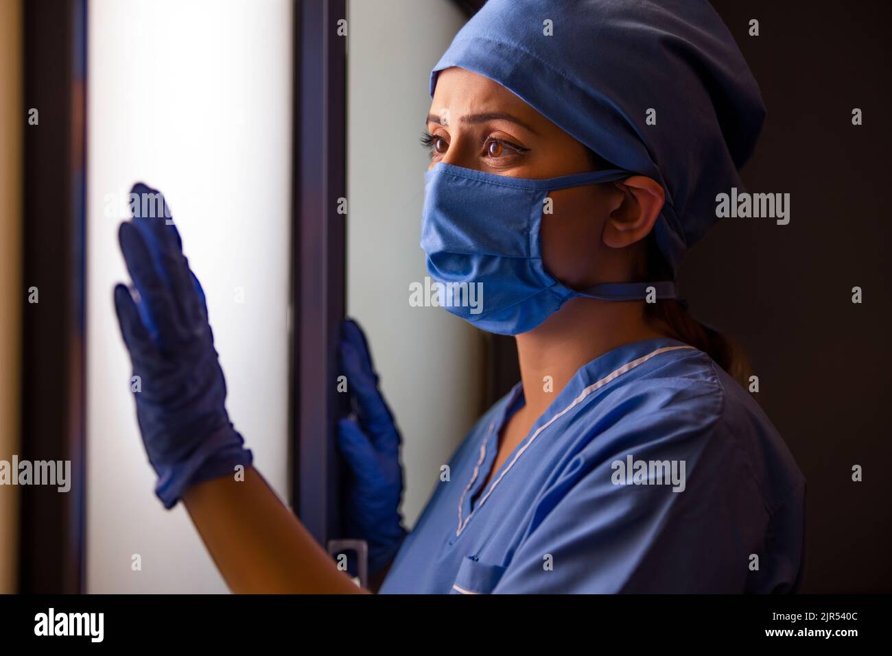Gros plan d'une jeune infirmière féminine réfléchie regardant par la fenêtre à l'hôpital. Banque D'Images Gros plan d'une jeune infirmière féminine réfléchie regardant par la fenêtre à l'hôpital. Banque D'Images