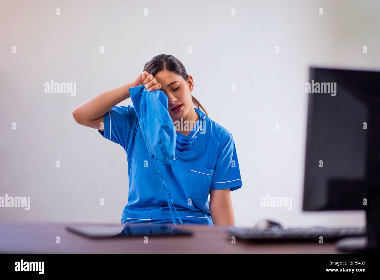 Portrait d'une infirmière fatiguée qui retire la coiffe chirurgicale tout en étant assise sur un bureau d'ordinateur Banque D'Images