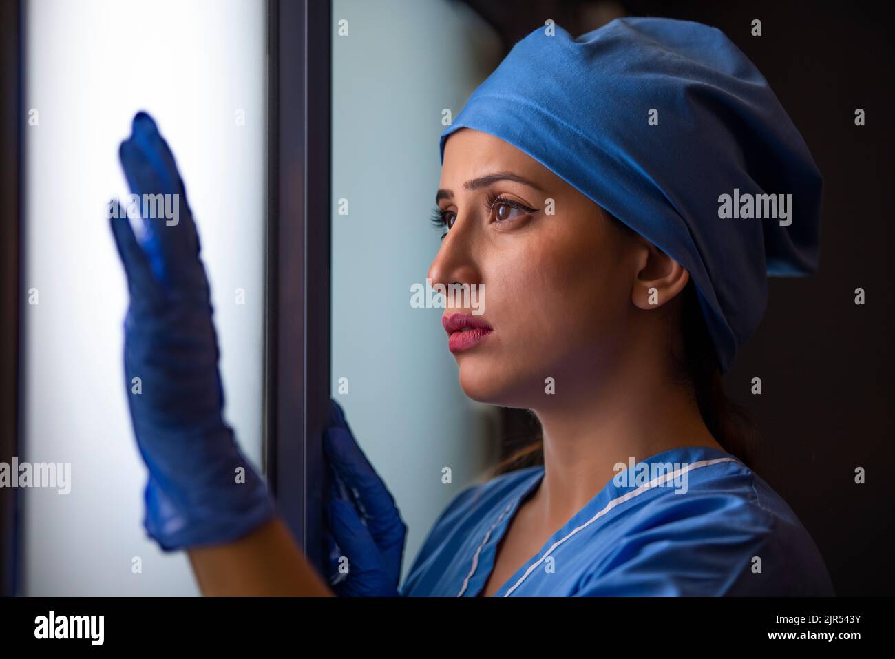 Gros plan d'une jeune infirmière féminine réfléchie regardant par la fenêtre à l'hôpital. Banque D'Images Gros plan d'une jeune infirmière féminine réfléchie regardant par la fenêtre à l'hôpital. Banque D'Images
