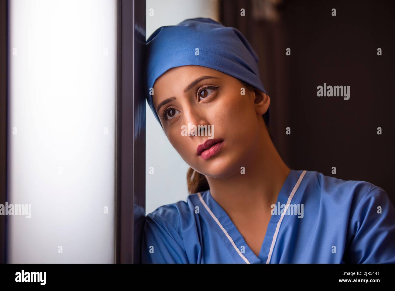 Gros plan d'une jeune infirmière féminine réfléchie regardant par la fenêtre à l'hôpital. Banque D'Images Gros plan d'une jeune infirmière féminine réfléchie regardant par la fenêtre à l'hôpital. Banque D'Images