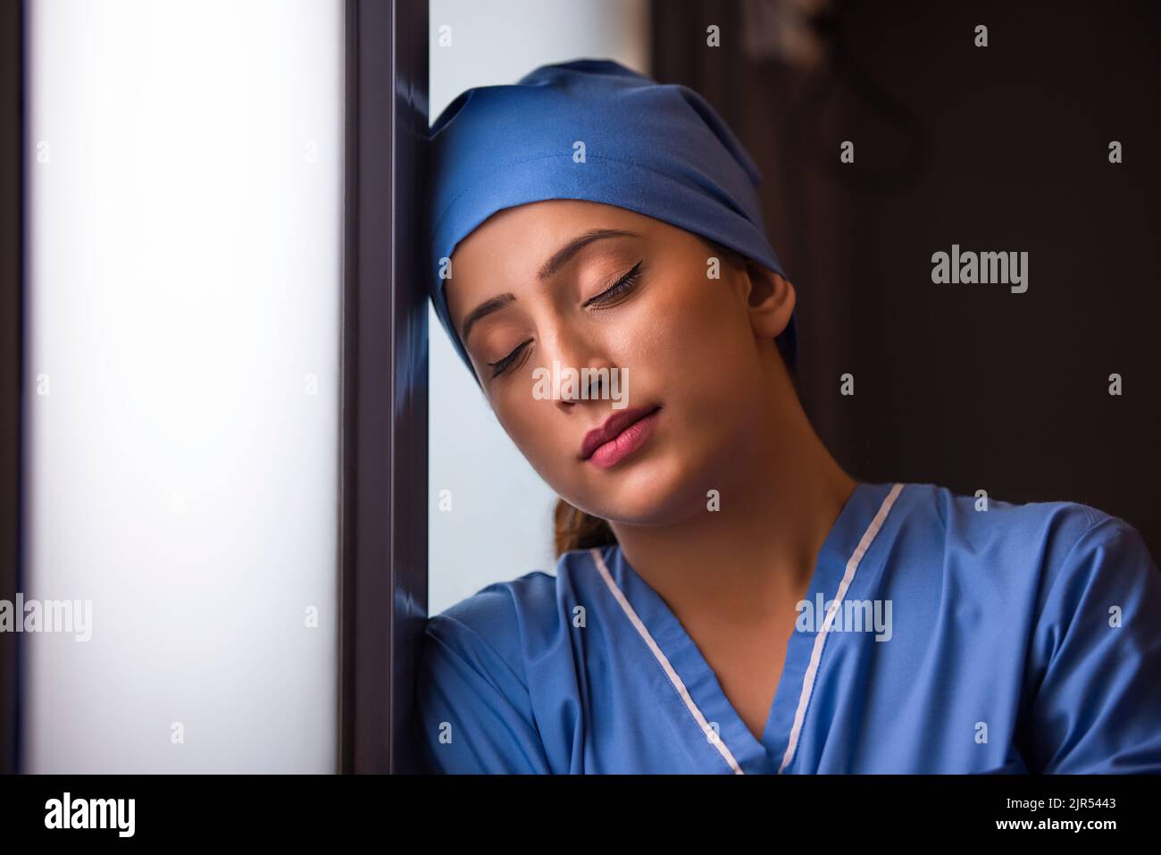 Gros plan d'une infirmière femme épuisée debout près de la fenêtre, les yeux fermés à l'hôpital. Banque D'Images Gros plan d'une infirmière femme épuisée debout près de la fenêtre, les yeux fermés à l'hôpital. Banque D'Images