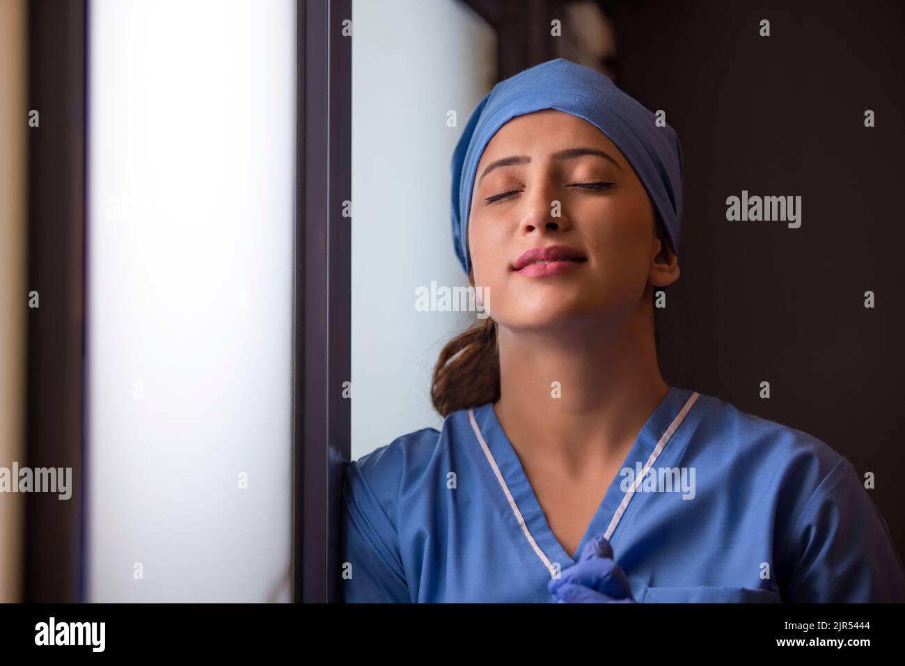 Gros plan d'une infirmière femme épuisée debout près de la fenêtre, les yeux fermés à l'hôpital. Banque D'Images Gros plan d'une infirmière femme épuisée debout près de la fenêtre, les yeux fermés à l'hôpital. Banque D'Images