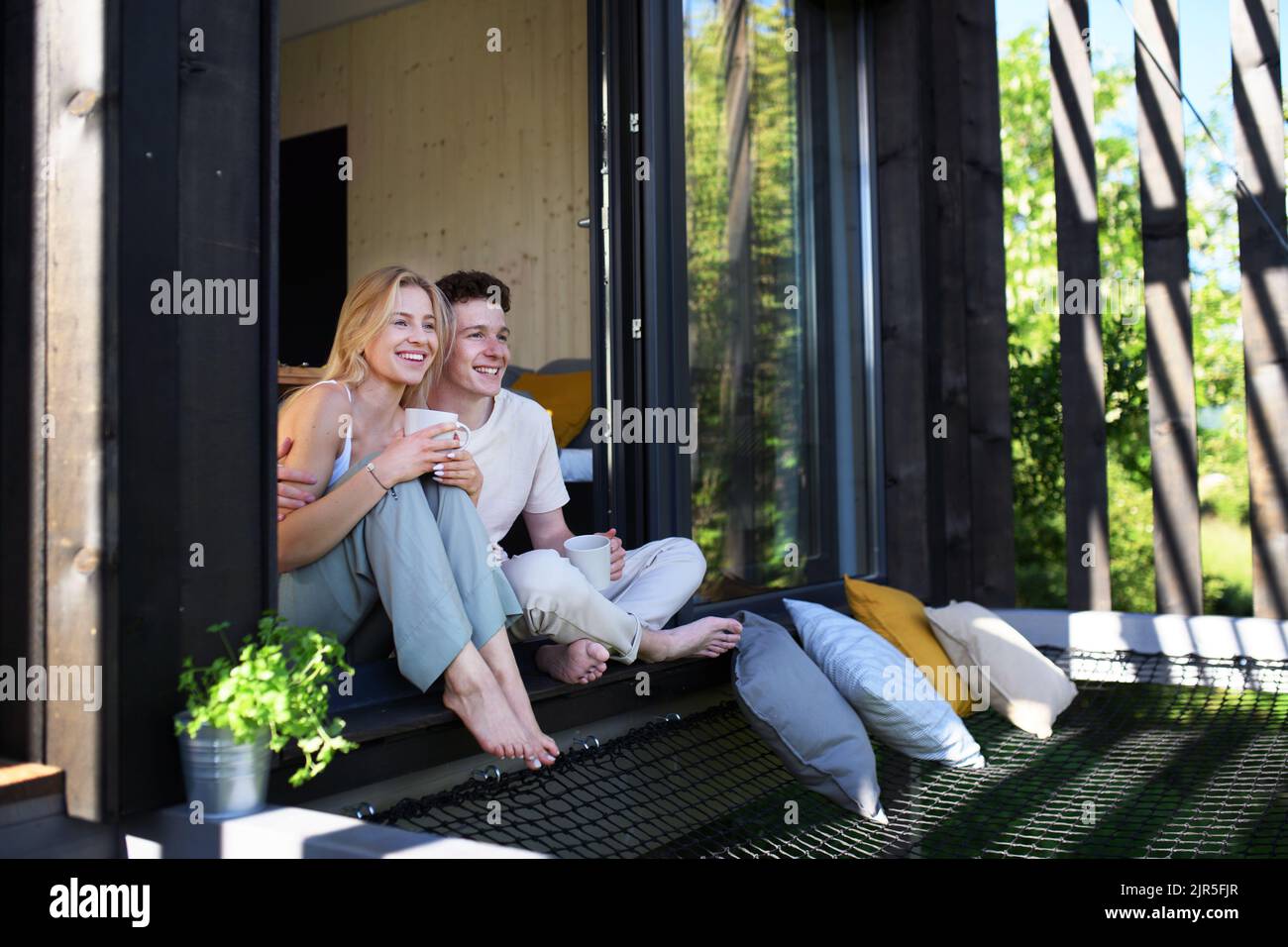 Jeune couple assis avec une tasse de café et en-cas en terrasse dans leur nouvelle maison dans une petite maison en bois, concept de vie durable. Banque D'Images