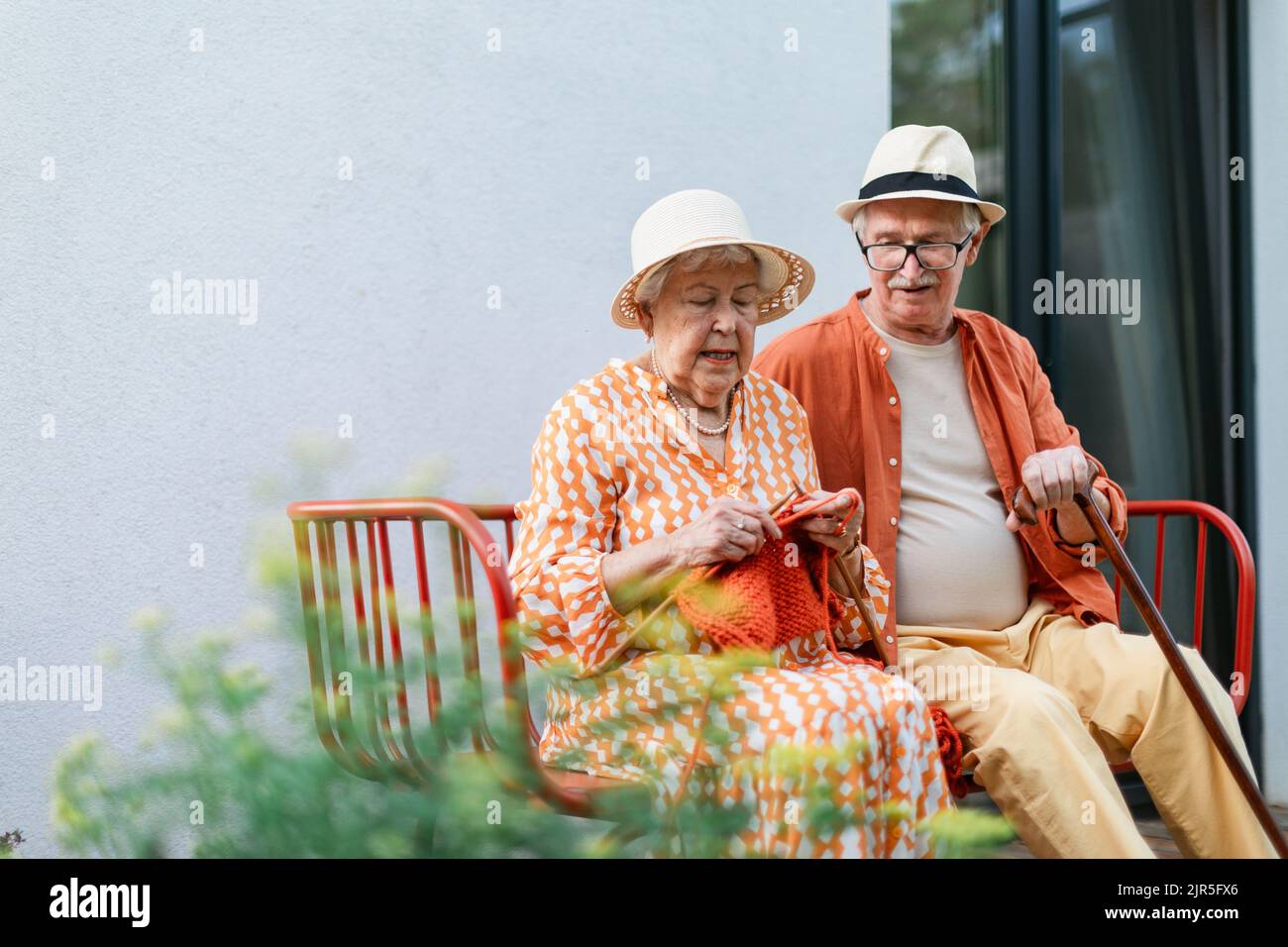 Femme âgée assise dans le jardin sur le banc avec son mari et tricoter le foulard rouge. Banque D'Images