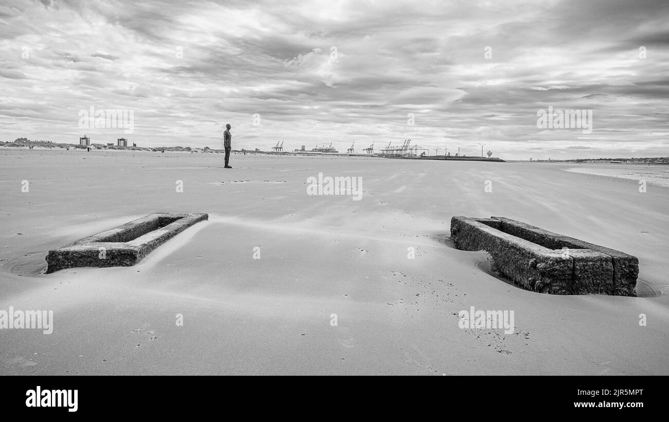 Une image en noir et blanc d'un Iron Man sur une plage venteuse de Crosby près de Liverpool vue en août 2022 derrière une paire de supports de tuyaux en bois. Banque D'Images