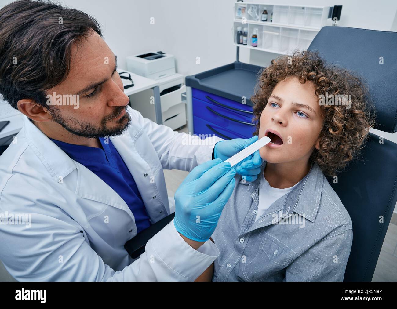 docteur utilisant la spatule d'inspection pour examiner la gorge bouclée du garçon. Enfant mâle au rendez-vous du pédiatre Banque D'Images