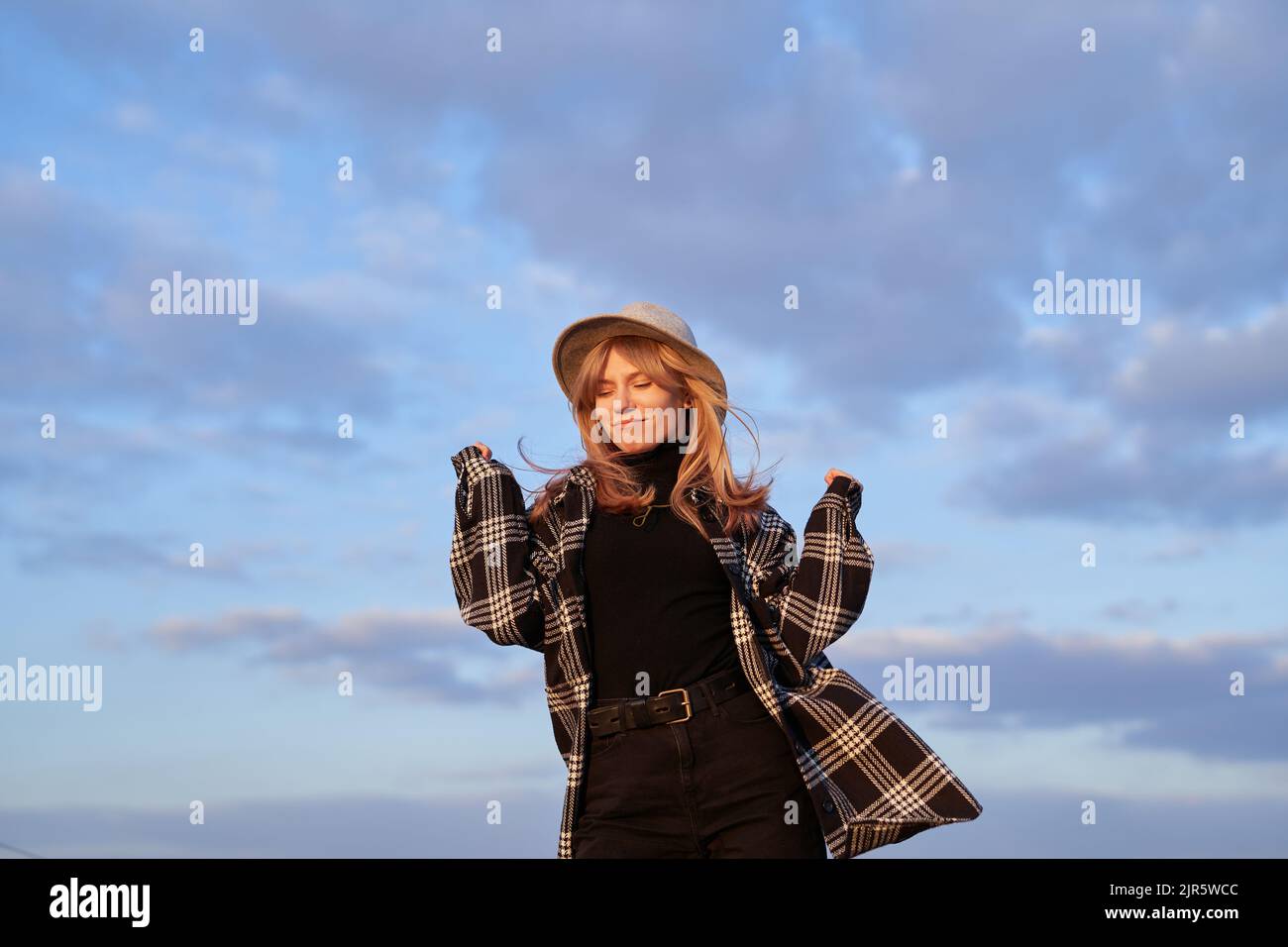 Concept gagnant, expressif ou positif. Jeune fille caucasienne en chapeau, jeans noirs et chemise dansant sensation joyeuse ou insouciante d'émotion devant le ciel bleu nuageux. Image de haute qualité Banque D'Images