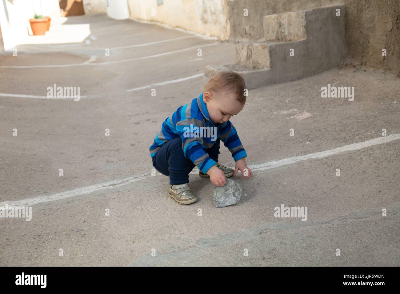 Bébé caucasien jouant à l'extérieur avec une pierre et un rocher dans la rue. Espagne. Série Banque D'Images