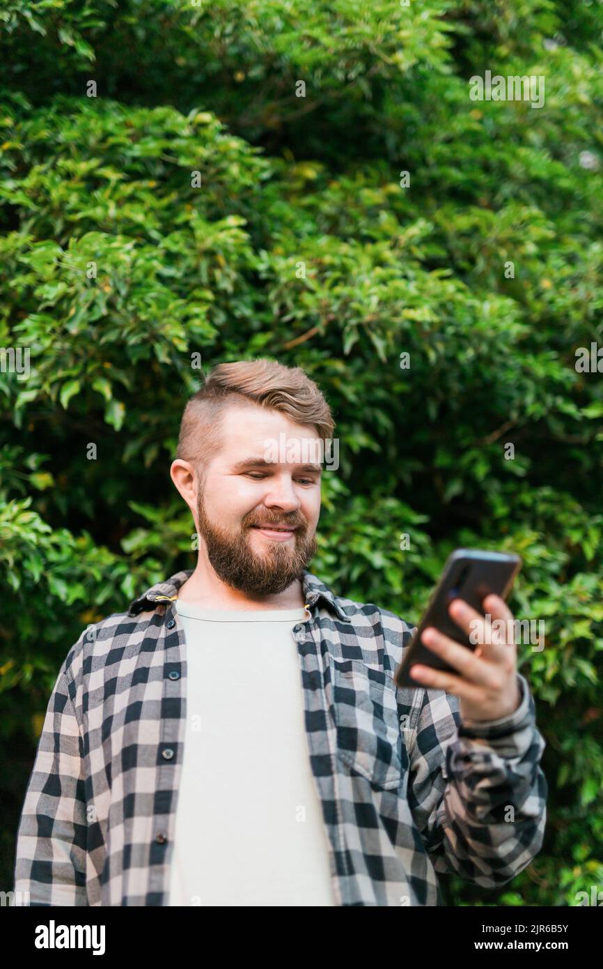 Portrait d'un homme joyeux et attrayant utilisant un smartphone pour faire défiler sur les réseaux sociaux sur fond d'arbre vert Banque D'Images