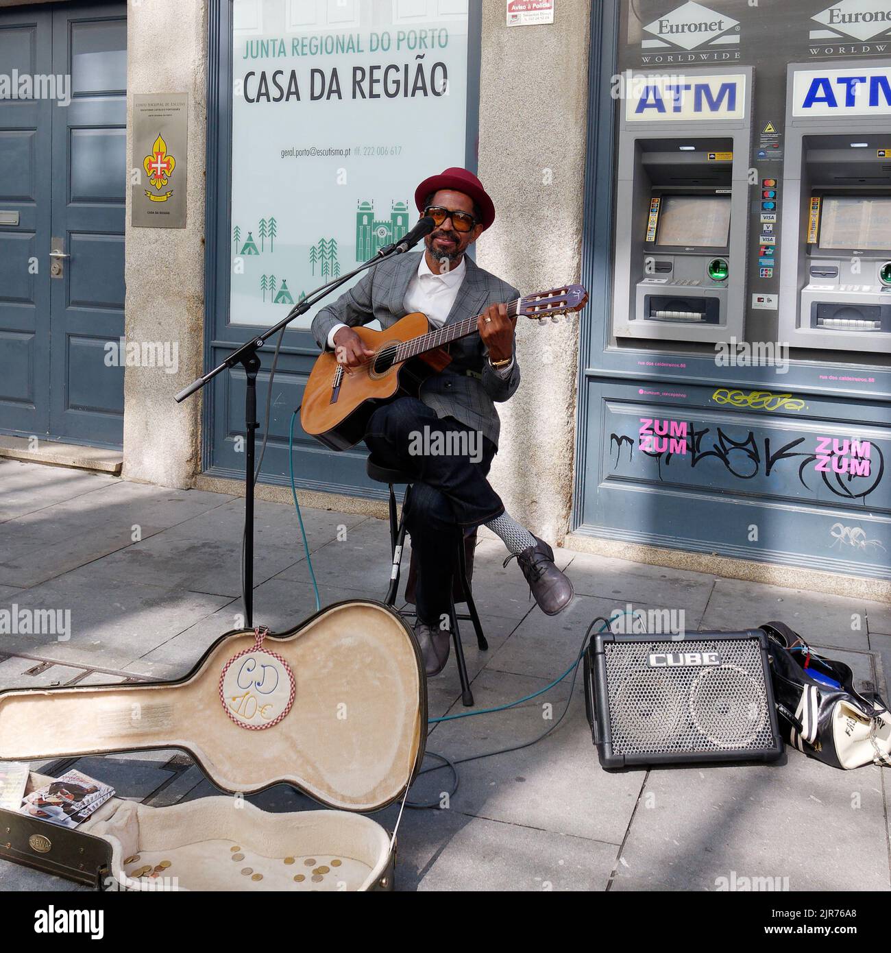 Busker portant un chapeau joue une guitare dans les rues de Porto, au Portugal, avec un haut-parleur et un étui de guitare devant et un distributeur automatique de billets derrière. Banque D'Images