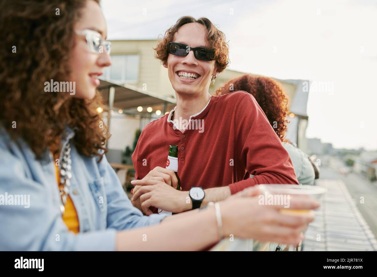 Jeune homme gai avec une bouteille de bière debout devant sa petite amie sur la terrasse du café sur le toit tout en appréciant la fête en plein air Banque D'Images