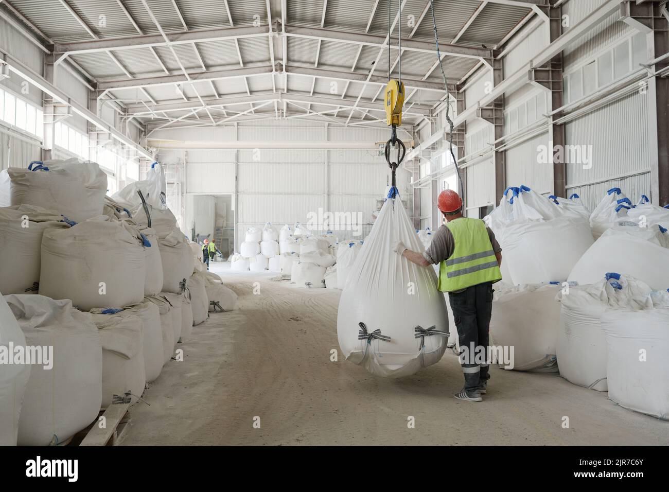 Vue arrière de jeune homme ouvrier de l'usine en uniforme et casque de sécurité se déplaçant le long des rangées d'énormes sacs lourds avec des matières premières en vrac Banque D'Images