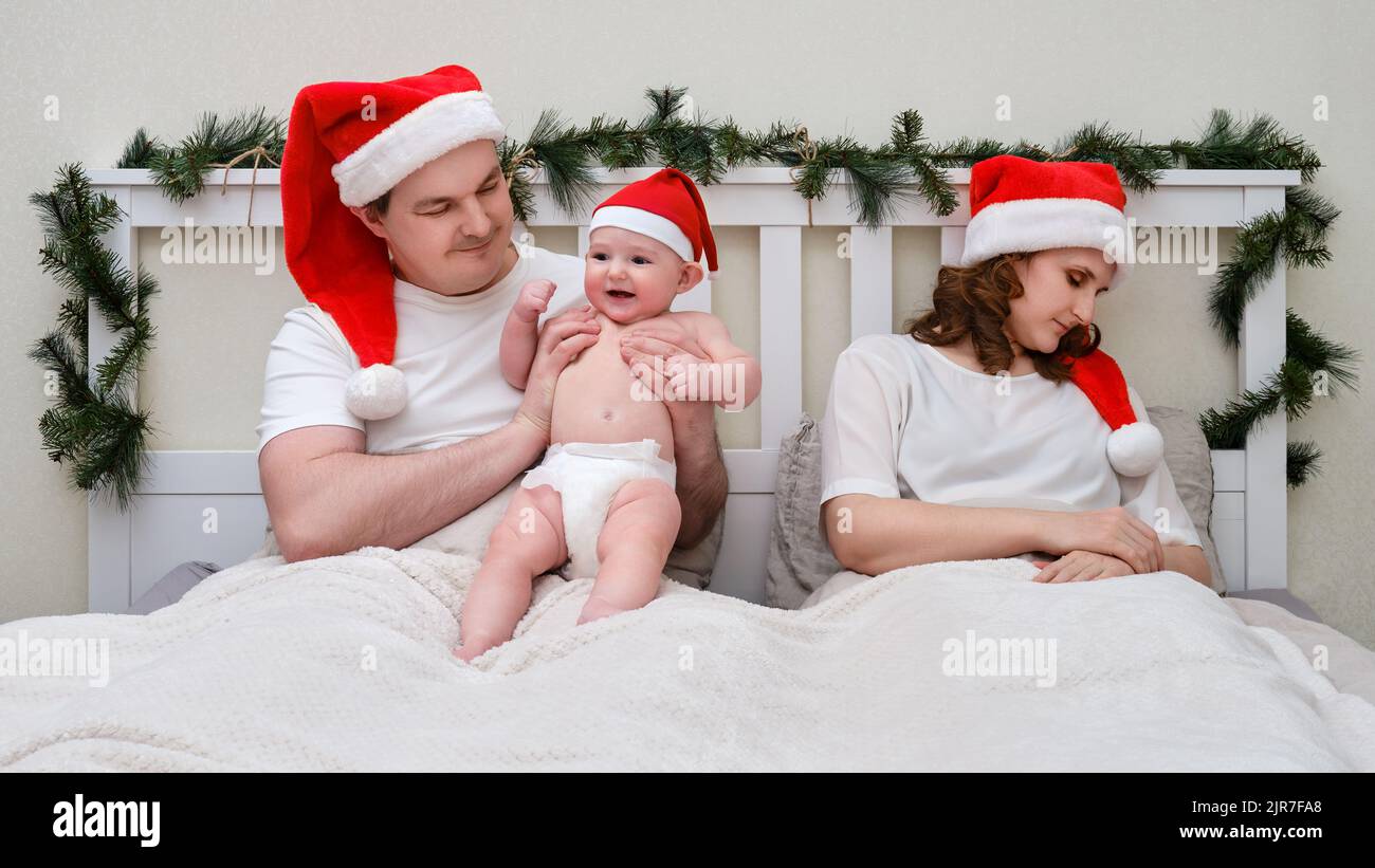 Le mari joue avec l'enfant tandis que la femme dort sur le lit décoré pour noël et nouvel an, couple de famille dans la chambre à la maison Banque D'Images