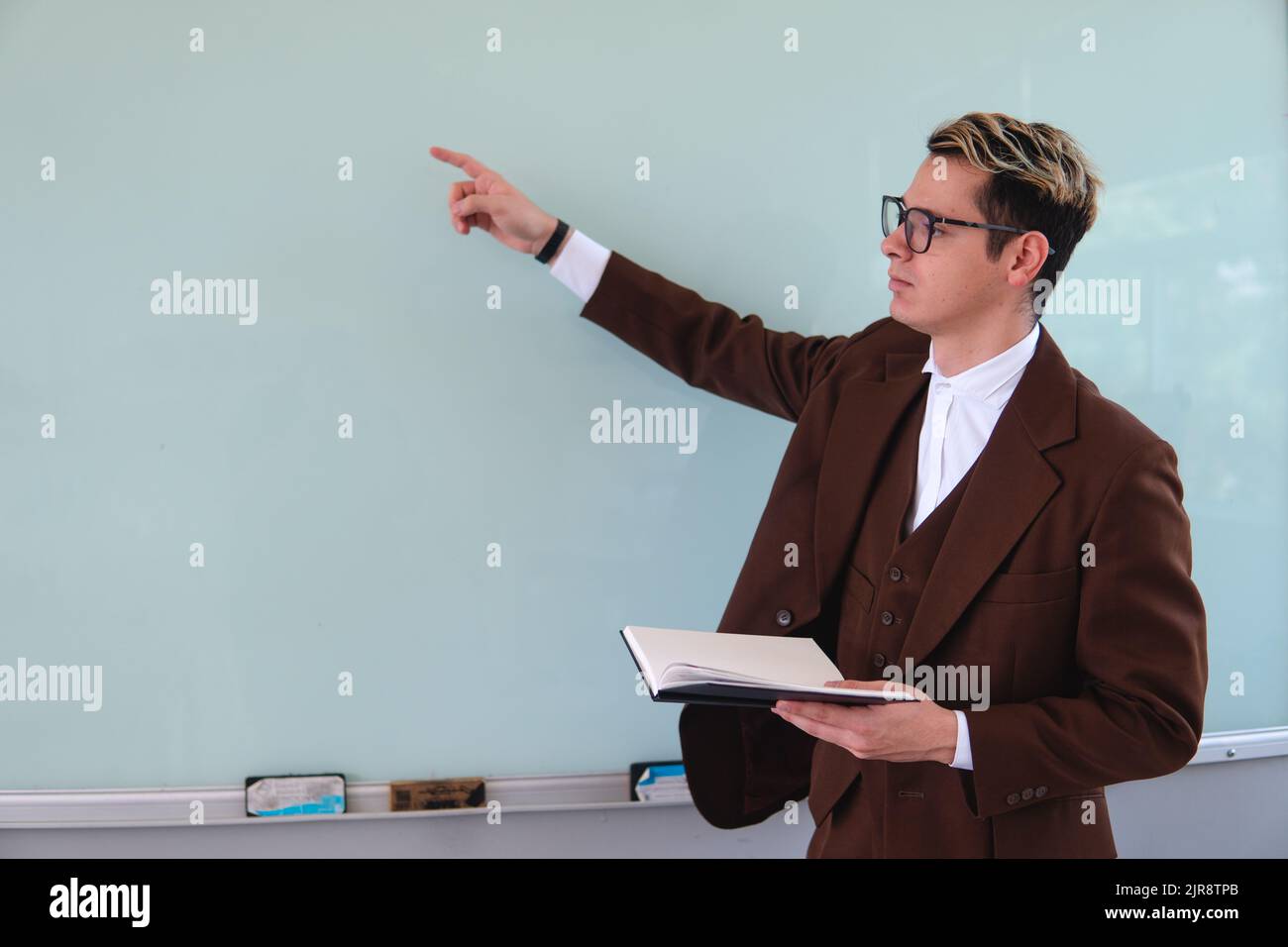 Un professeur pointant vers un tableau noir. Professeur d'université avec un livre noir dans sa main. Maître habillé dans un élégant costume brun. Homme élégant dans un tableau noir expliquant un sujet. Photo de haute qualité Banque D'Images