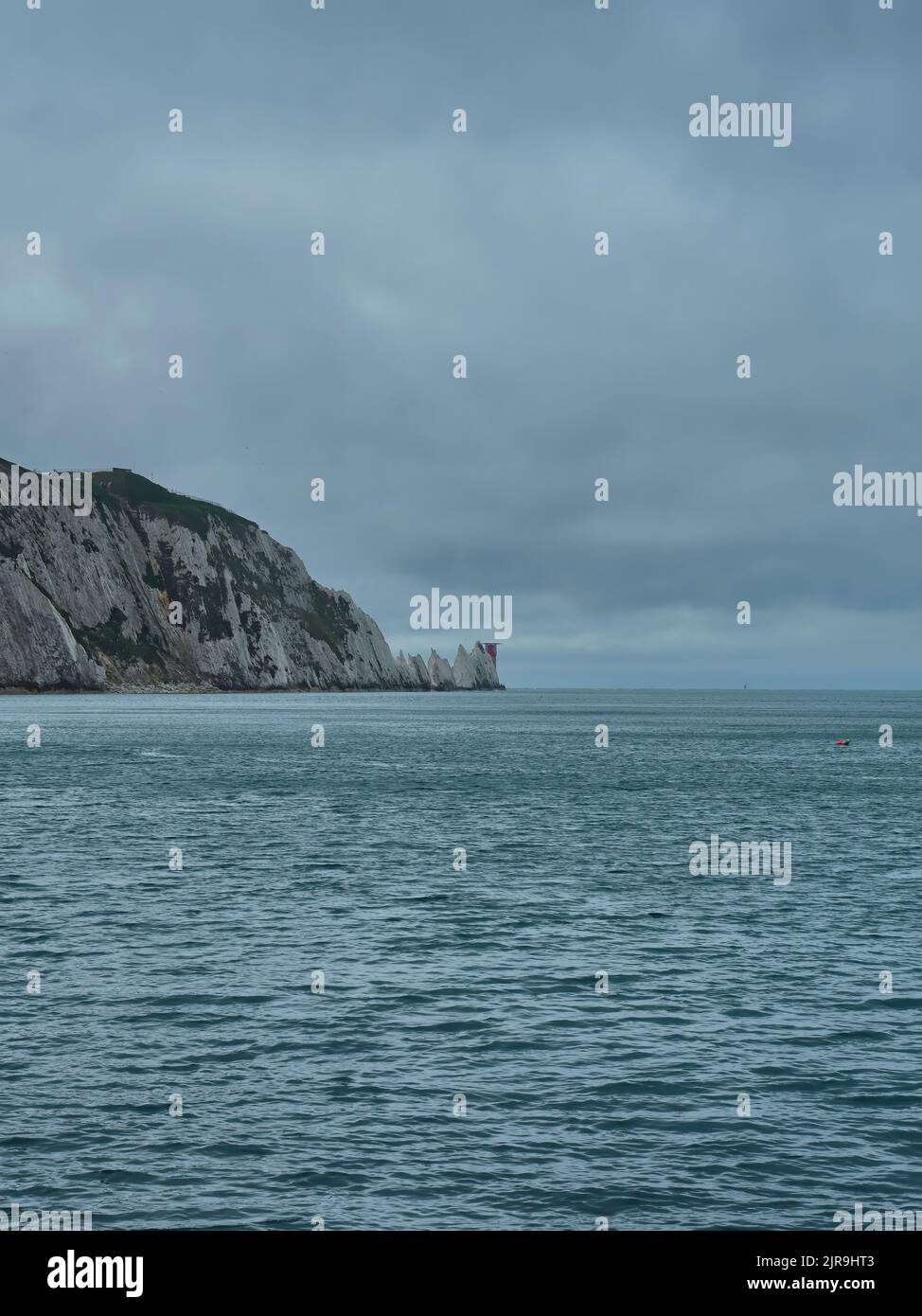Le phare au niveau des aiguilles derrière des affleurements dentelés, qui expliquent clairement pourquoi le phare est nécessaire, dans une mer à ciel ouvert sous un ciel couvert. Banque D'Images