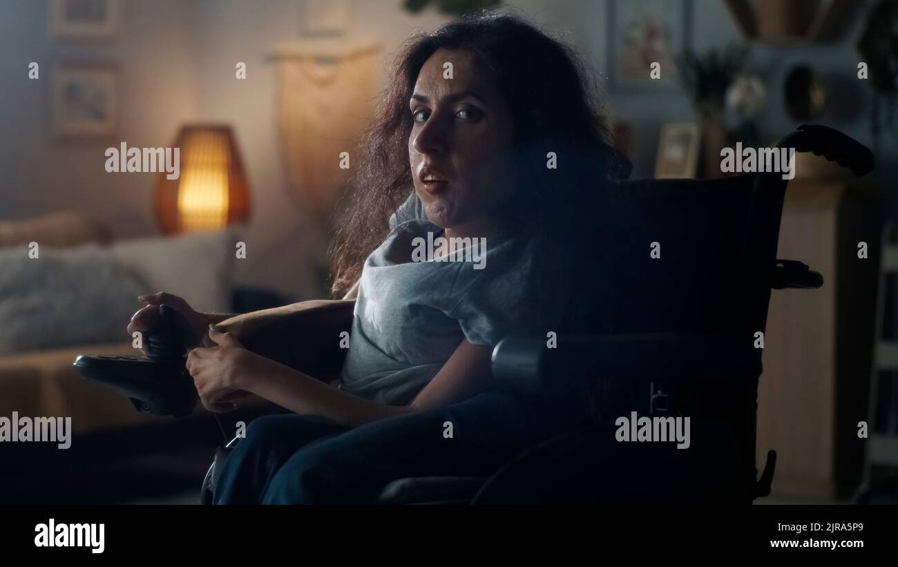 Triste femme handicapée entrant dans la pièce sombre de la maison confortable et regardant la caméra tout en étant assise dans un fauteuil roulant motorisé Banque D'Images