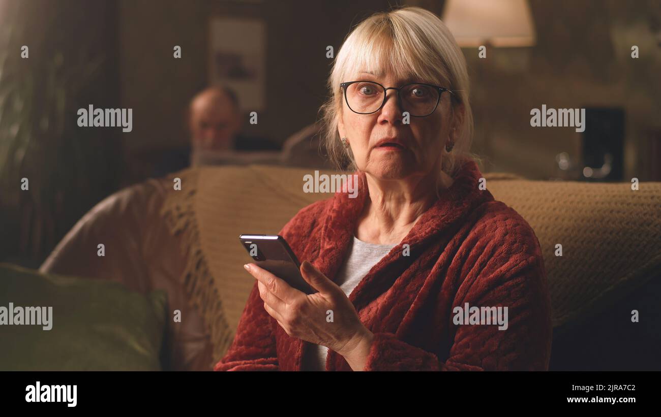 Une femme effrayée dans des lunettes regardant un appareil photo à la bouche ouverte, assis sur un canapé après avoir lu des nouvelles choquantes sur un téléphone portable le soir à la maison Banque D'Images