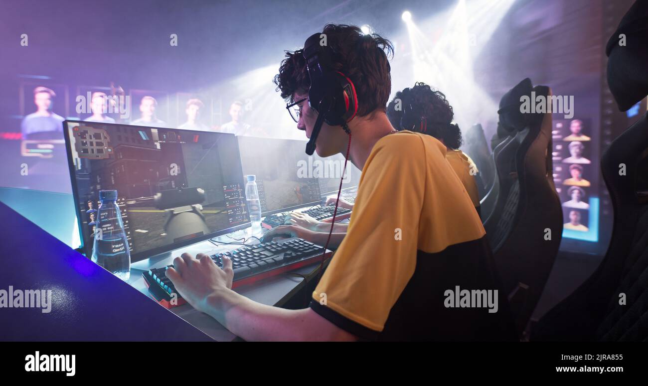 Joueur d'Esportsman dans des lunettes et des écouteurs jouant au tireur sur ordinateur près de l'équipe tout en prenant le parc dans le championnat de jeu professionnel Banque D'Images