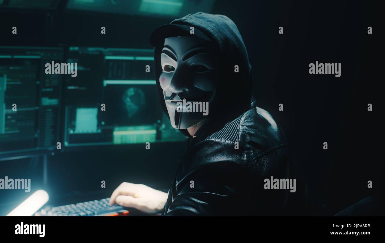 Un hacker dans un masque anonyme regardant la caméra pendant un cybercrime avec une pièce sombre avec des moniteurs et des équipements modernes Banque D'Images