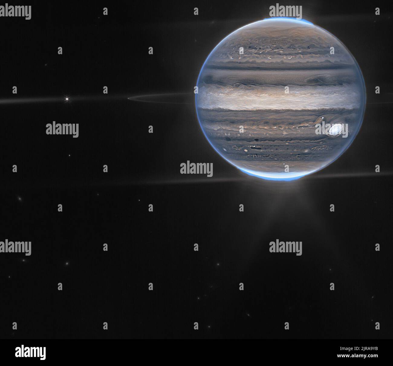 Description de l'image : une vue de champ large affiche Jupiter dans le quadrant supérieur droit. Les bandes horizontales tourbillonnantes de la planète sont rendues dans les bleus, les bruns et la crème. Auroras bleu électrique au-dessus des pôles nord et sud de Jupiter. Une lueur blanche émane des aurores. Le long de l'équateur de la planète, les anneaux brillent dans un blanc pâle. Ces anneaux sont un million de fois plus faibles que la planète elle-même! À l'extrémité gauche des anneaux, une lune apparaît sous la forme d'un petit point blanc. Légèrement plus à gauche, une autre lune brille avec de minuscules pics de diffraction blancs. Le reste de l'image est la noirceur de sp Banque D'Images