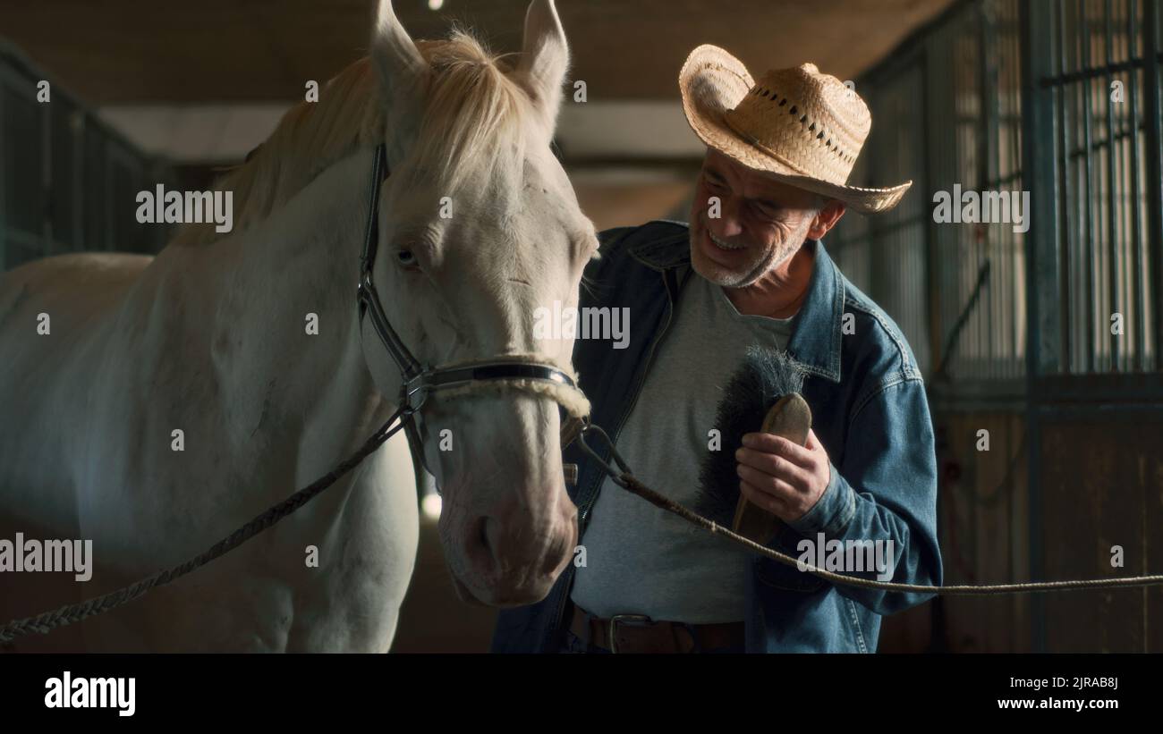 Homme âgé en chapeau et veste en denim éliminant la saleté du manteau de cheval albinos dans l'allée de la grange près des étals dans la journée à la ferme Banque D'Images
