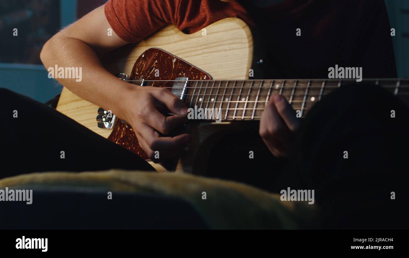 Inclinez la vue de l'adolescent concentré assis sur le lit et jouant de la guitare électrique pendant la séance de musique dans une chambre obscure la nuit à la maison Banque D'Images