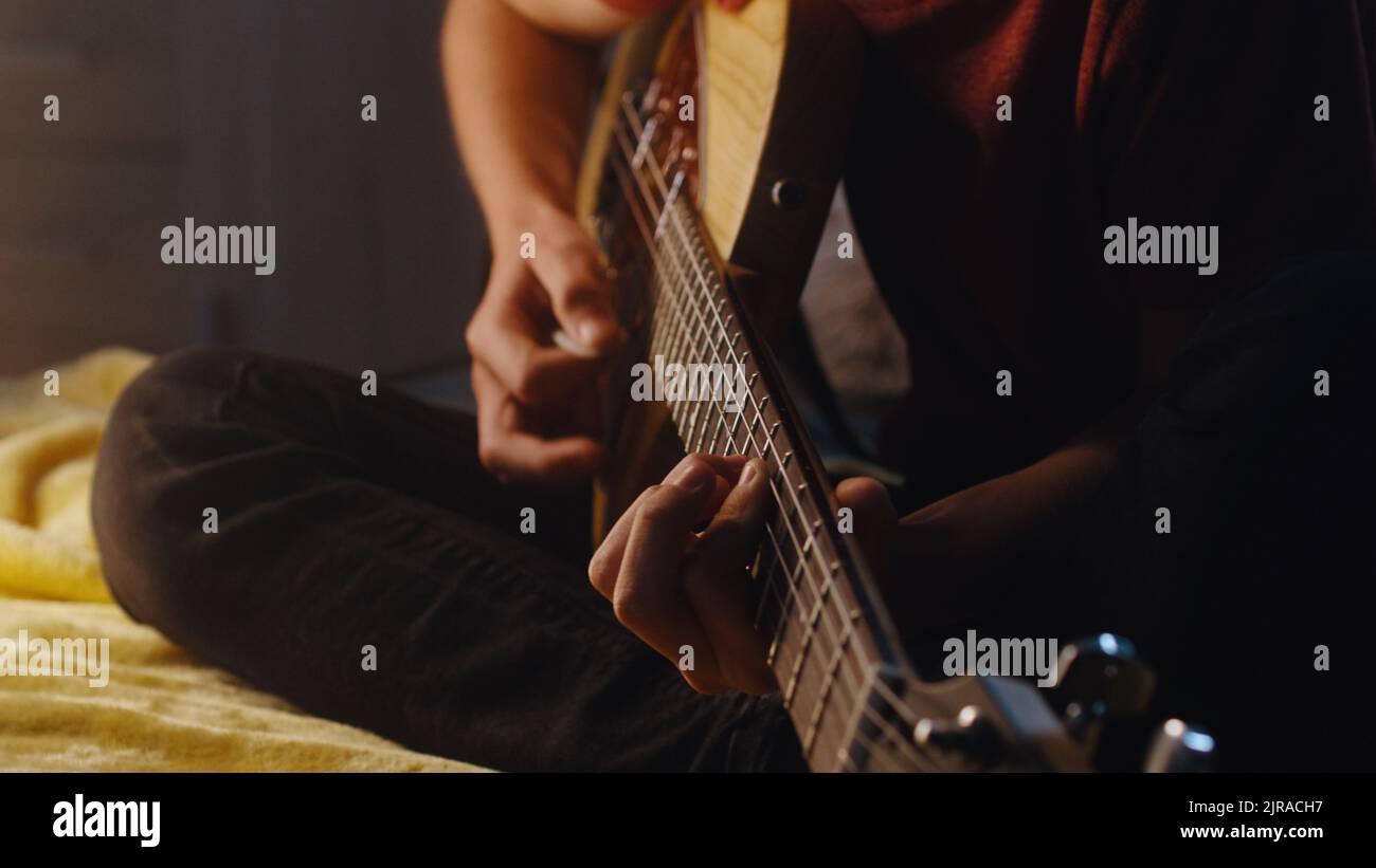 Inclinez la vue de l'adolescent concentré assis sur le lit et jouant de la guitare électrique pendant la séance de musique dans une chambre obscure la nuit à la maison Banque D'Images