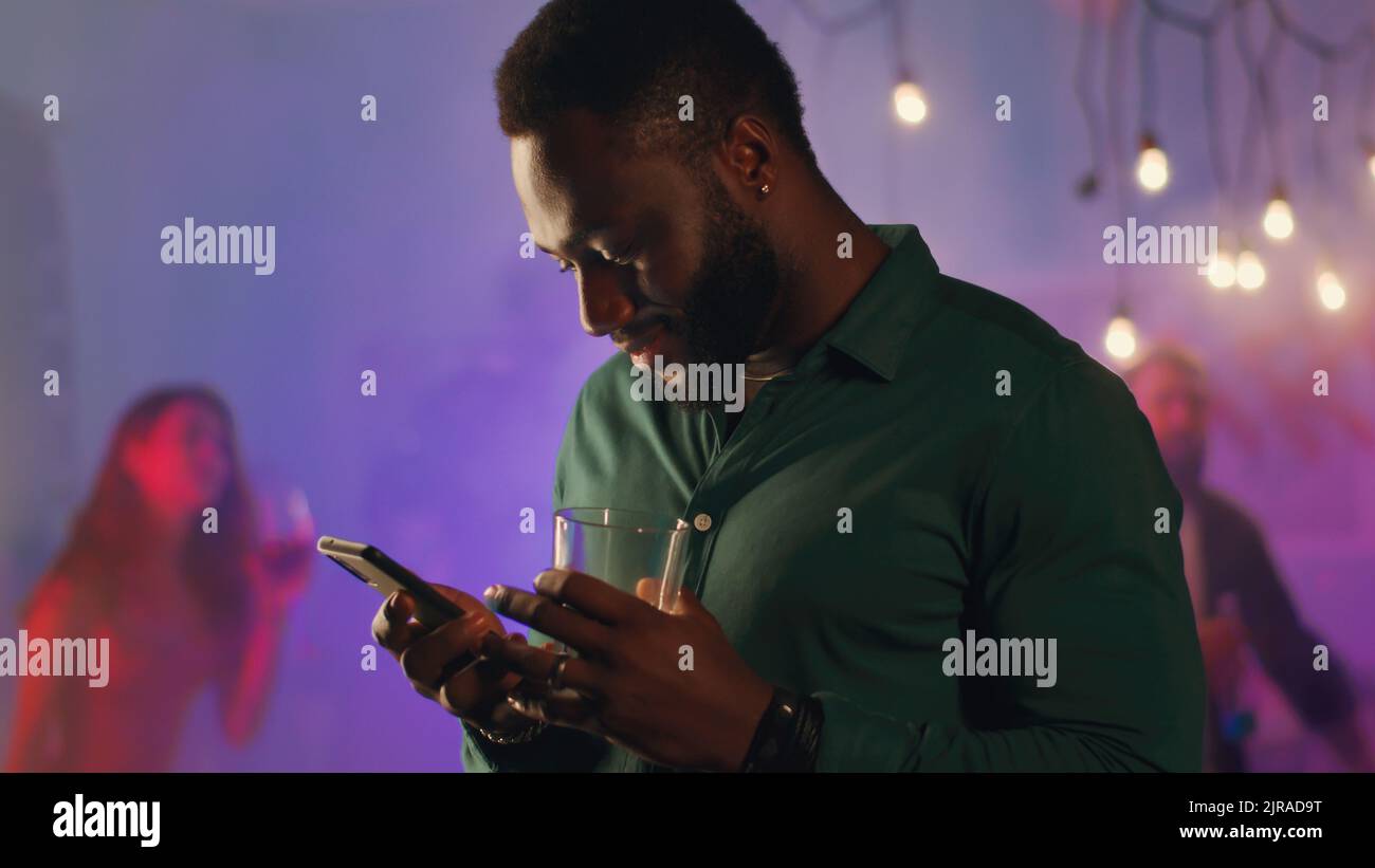Optimiste Africain américain homme avec l'alcool de queue de colecture et d'envoi de messages texte sur le téléphone portable pendant la fête à la maison dans la pièce remplie de fumée Banque D'Images