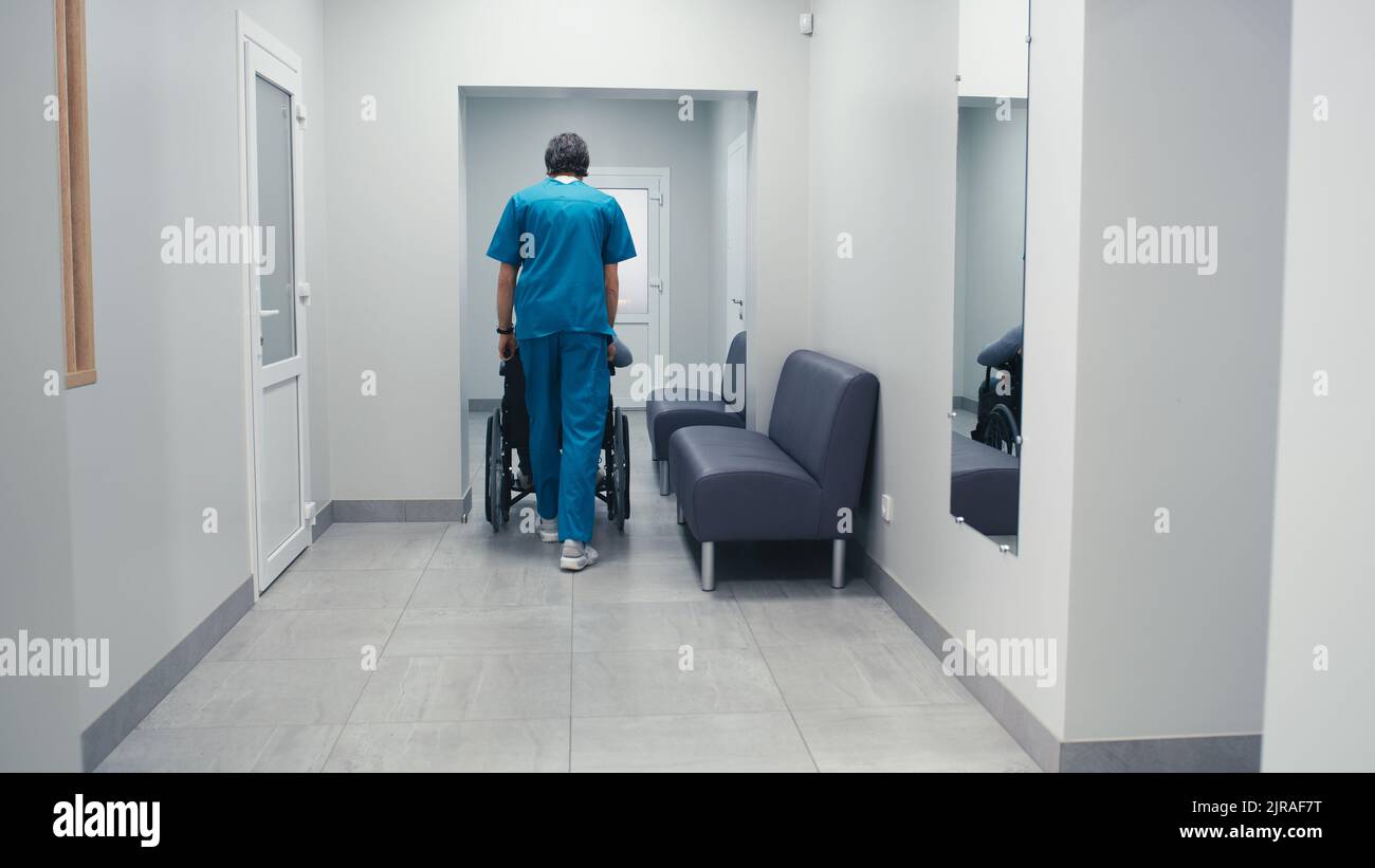 Vue arrière de l'homme dans les exfoliants poussant le fauteuil roulant avec un homme handicapé patient tout en travaillant dans le couloir de la clinique moderne Banque D'Images