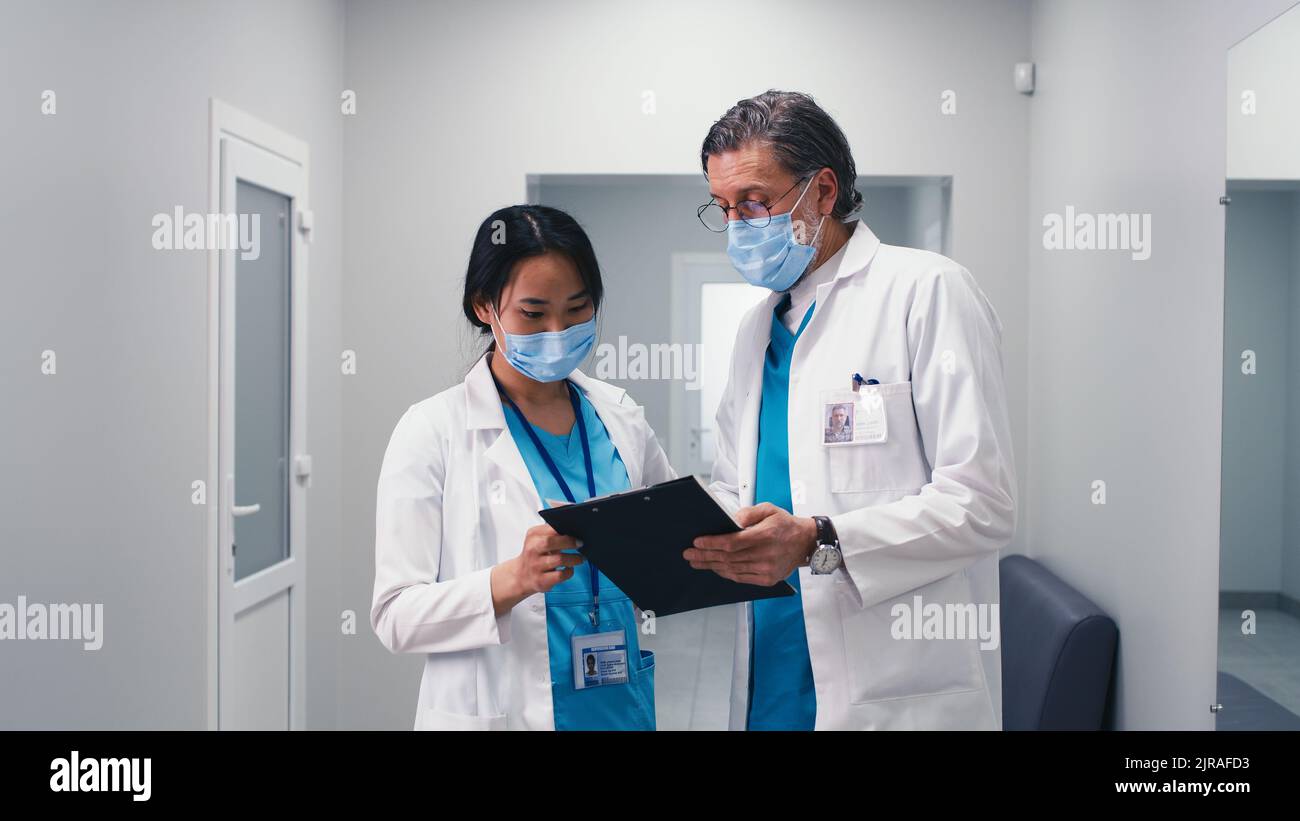 Homme adulte dans le masque discutant des données sur le presse-papiers avec une collègue asiatique puis s'en éloignant pendant le travail dans un centre médical Banque D'Images