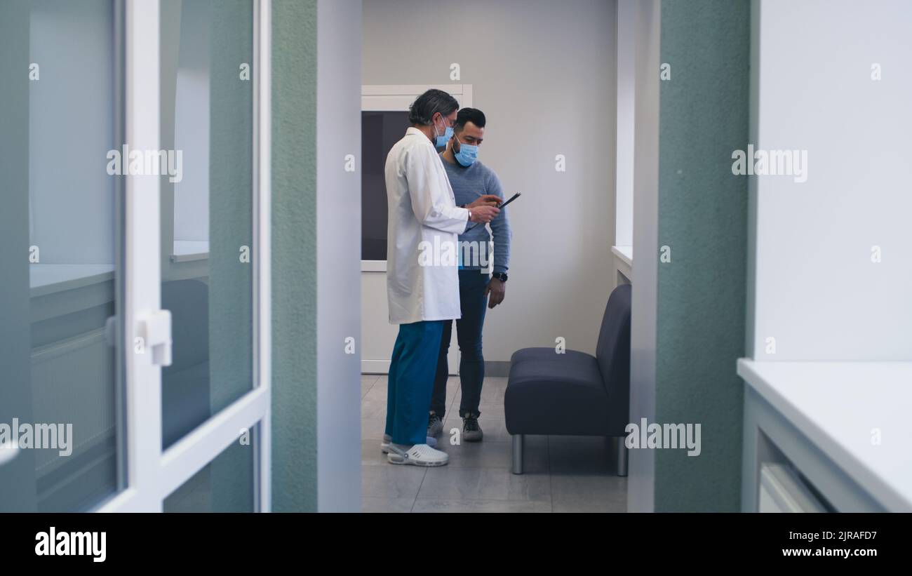 Homme anxieux en masque debout et à l'écoute d'un médecin d'âge moyen dans le couloir de l'hôpital moderne Banque D'Images