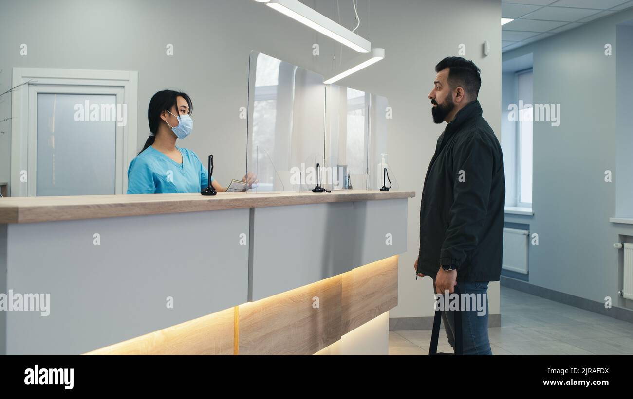 Homme barbu dans des vêtements décontractés parlant avec une réceptionniste asiatique en masque et un médecin homme mature lors d'une visite à l'hôpital moderne Banque D'Images
