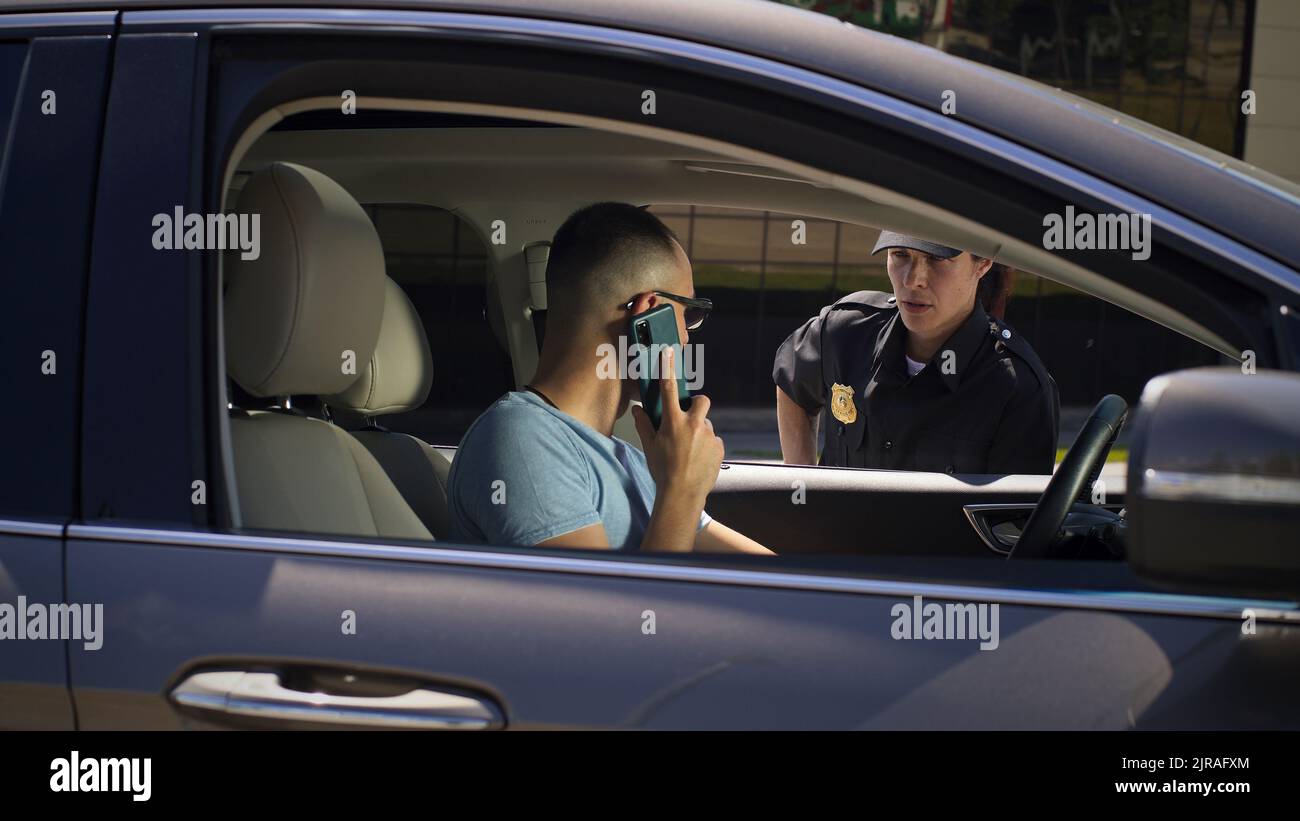 Homme en colère finissant l'appel téléphonique puis parlant et montrant le permis de conduire à une femme sérieuse en uniforme de police tout en étant assis dans la voiture dans la rue de la ville Banque D'Images