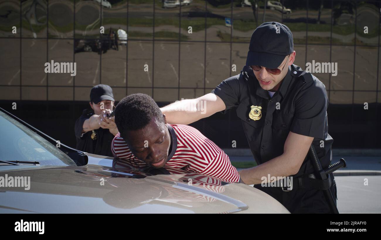 Homme et femme en uniforme de police visant le pistolet et la recherche résistant Afro-américain gars près de la voiture pendant l'arrestation Banque D'Images