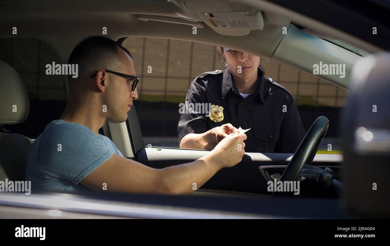 Femme sérieuse dans le permis de contrôle d'uniforme de police et de poser des questions à l'homme assis dans la voiture sur la rue de ville Banque D'Images