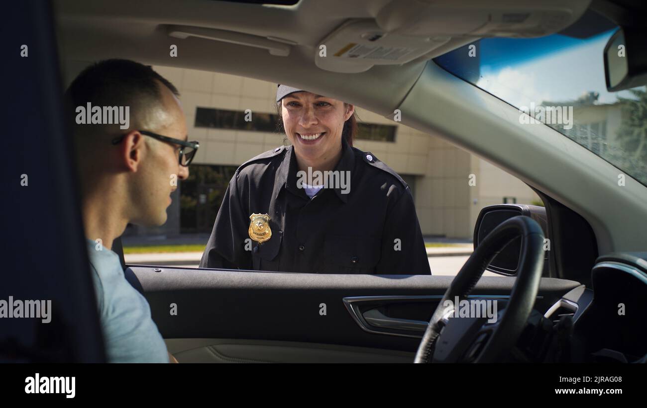 Femme gaie en uniforme de police sympathique souriant et vérifiant les documents de l'homme assis en voiture dans la rue de la ville Banque D'Images