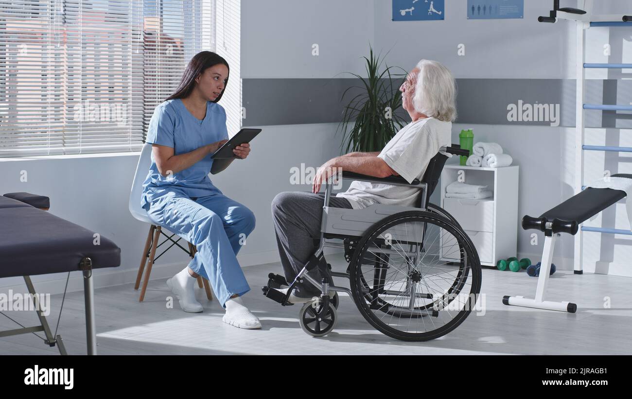 Homme âgé handicapé assis en fauteuil roulant et parlant avec une femme thérapeute en réadaptation à la clinique Banque D'Images