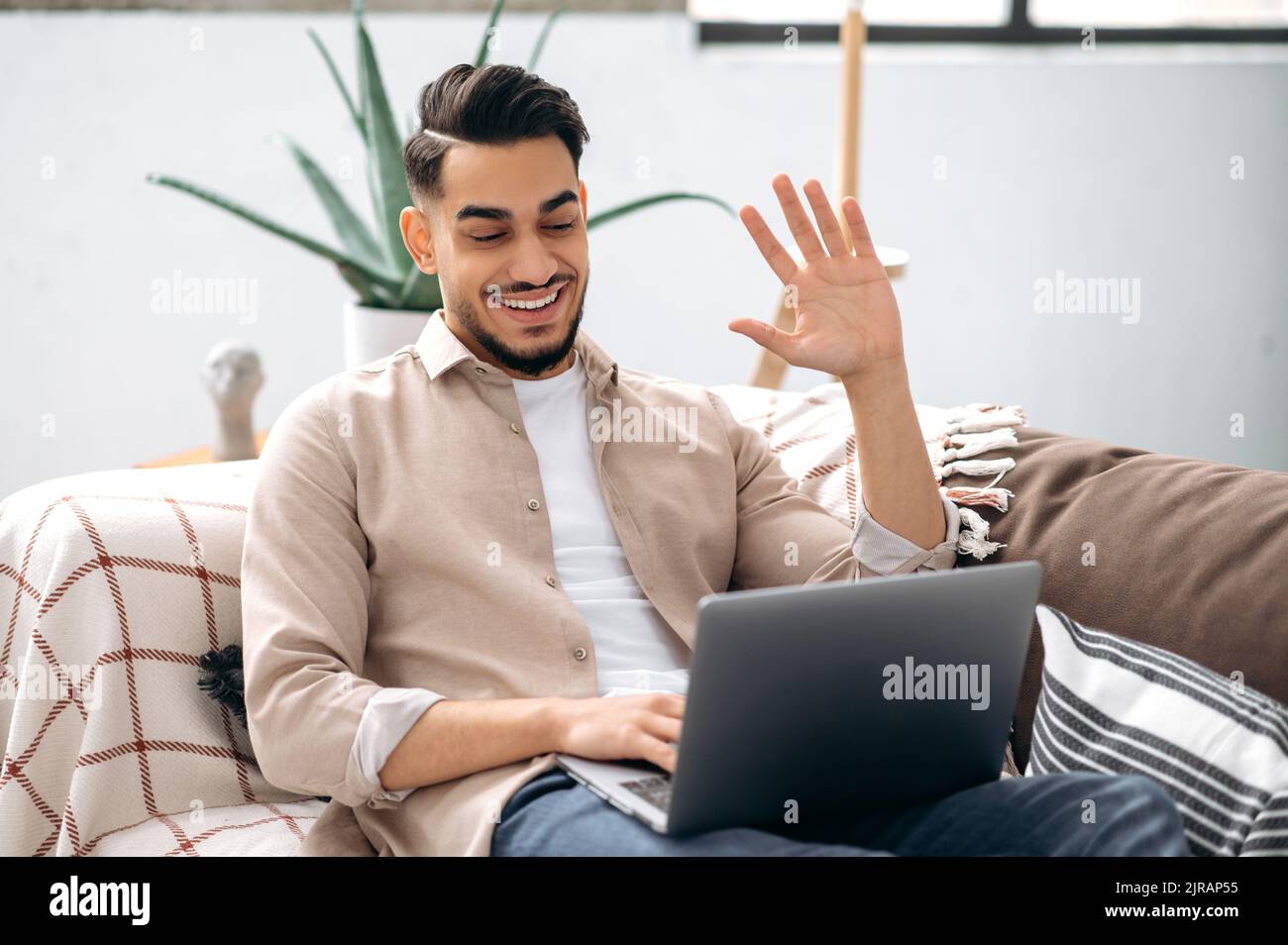 Communication vidéo en ligne. Jeune homme indien ou arabe sympathique, indépendant ou étudiant, assis sur un canapé à la maison dans la salle de séjour, parlant sur vidéo conférence avec un ami, collègue, client, sourires Banque D'Images