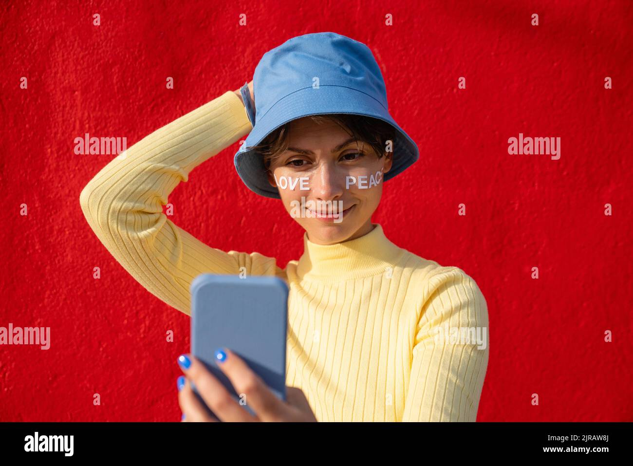 Femme avec un chapeau de seau prenant le selfie sur un smartphone devant le mur rouge Banque D'Images