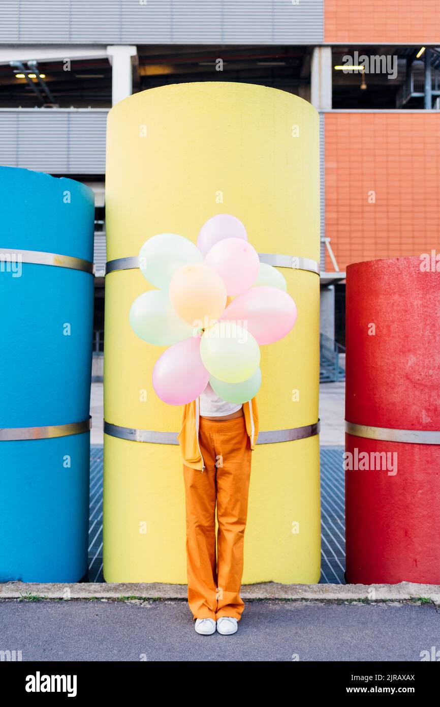 Jeune femme couvrant le visage avec des ballons colorés devant des tuyaux Banque D'Images