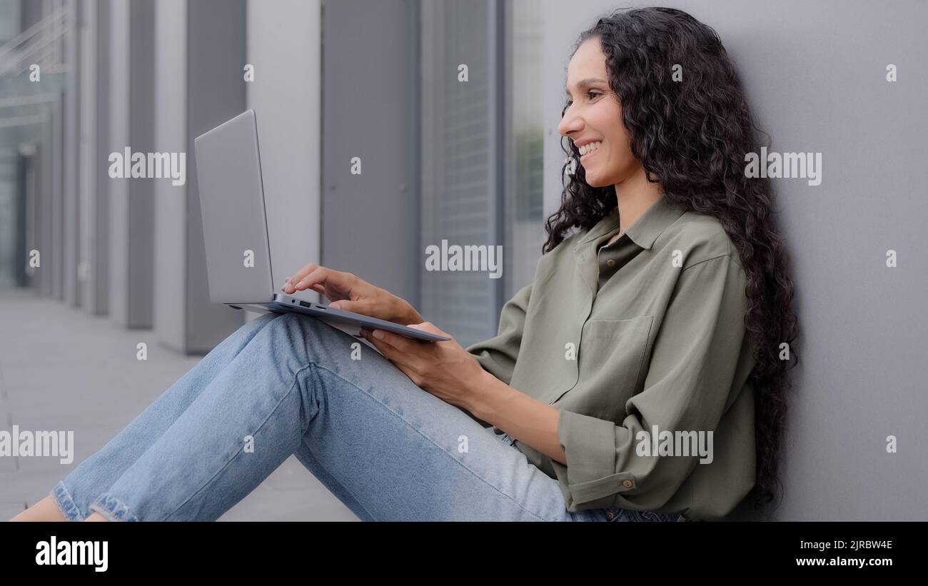 Souriante dame heureuse avec de longs cheveux assis sur le sol dactylographie navigation ordinateur portable décontracté chat femme utilisateur travailleur indépendant femme d'affaires penchée mur Banque D'Images