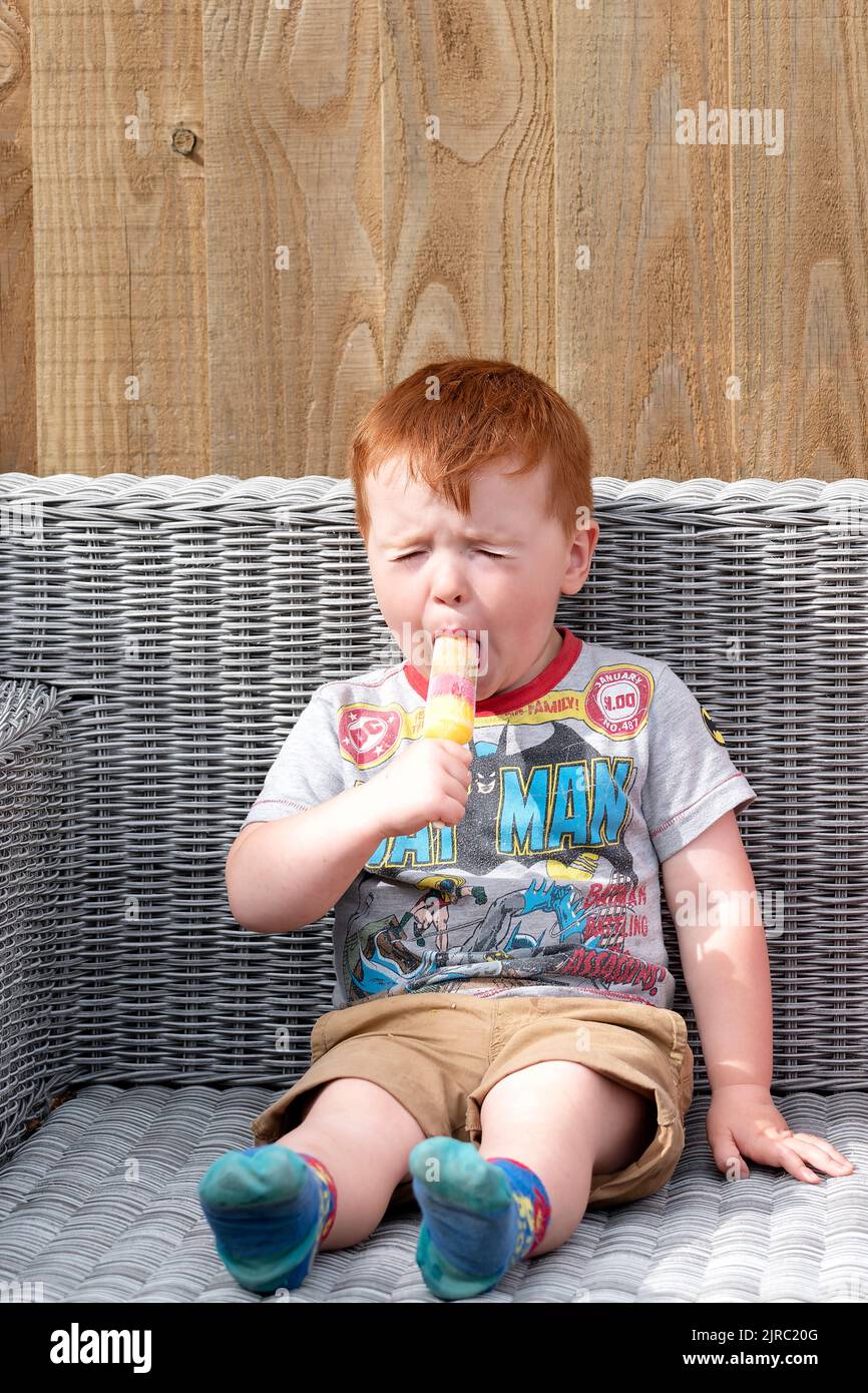 On montre à un jeune enfant de lécher un encloche de glace à saveur aigre. Le garçon est assis dehors et grimace le goût amer de la glace aromatisée. Banque D'Images