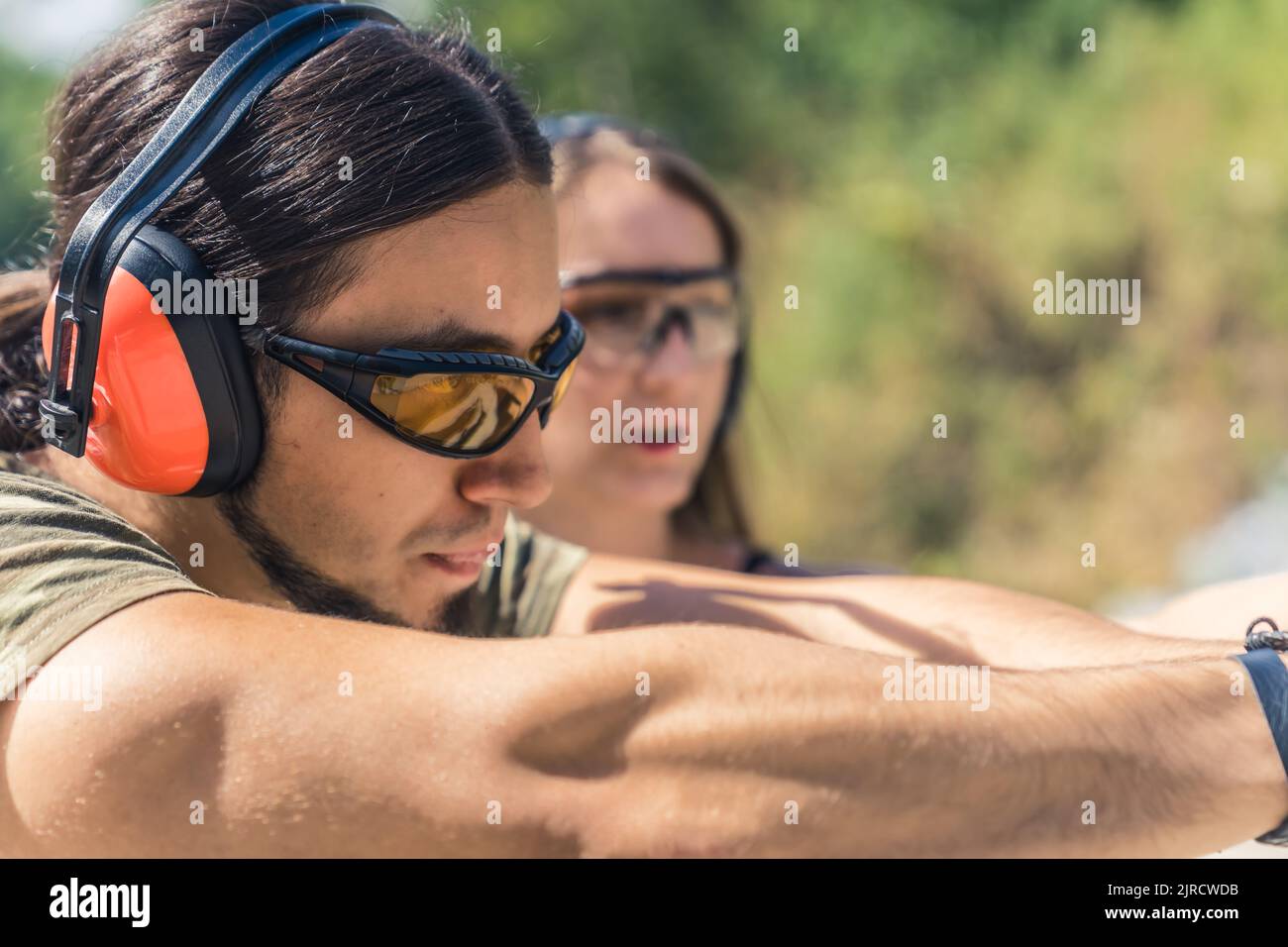 Gros plan d'un homme de race blanche portant des lunettes de sécurité et des écouteurs visant le pistolet observé par un instructeur féminin. Prise de vue horizontale en extérieur. Photo de haute qualité Banque D'Images