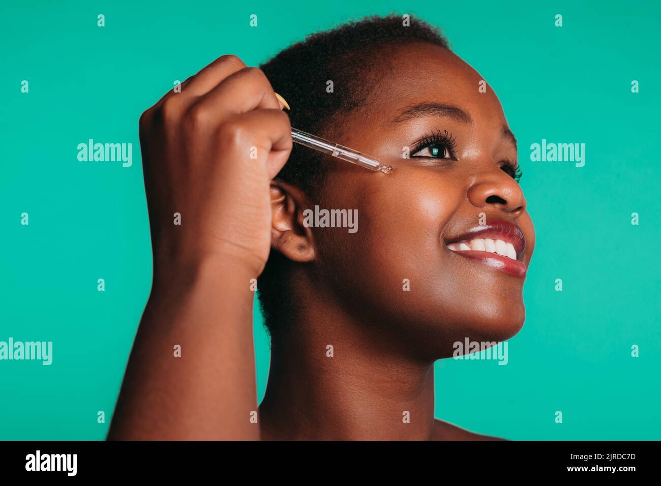 Portrait d'une jeune femme afro-américaine souriante appliquant du sérum facial avec compte-gouttes sur le visage, isolée sur fond turquoise. Sérum, huile pour le visage Banque D'Images