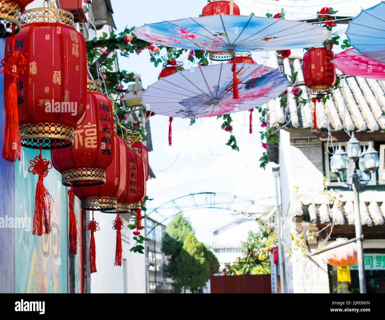Rue décorée avec des lanternes traditionnelles chinoises orientales rouges avec des rubans et des parasols ouverts à l'envers près d'un mur peint avec un dessin. Banque D'Images