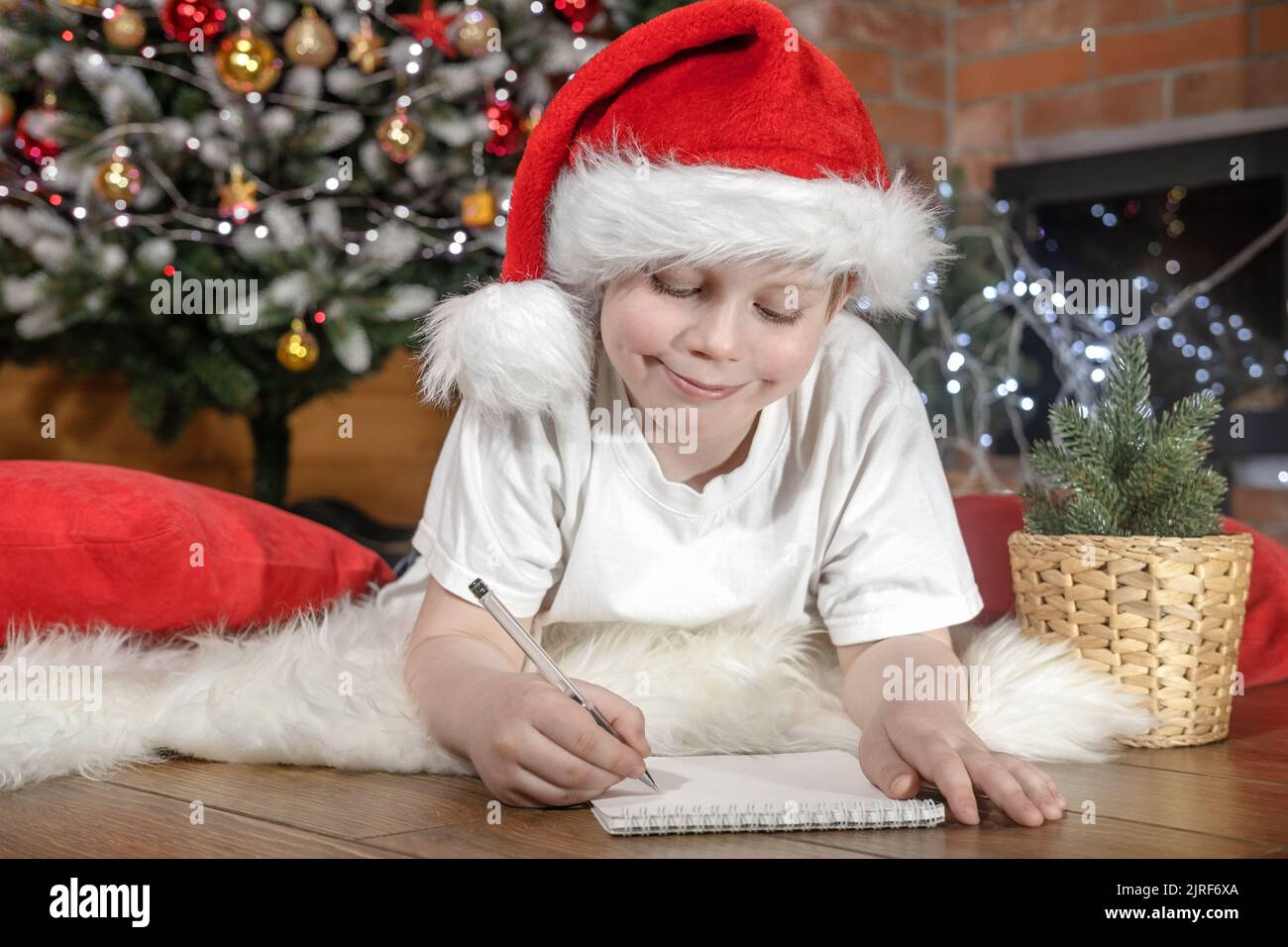Joyeux Noël aux enfants. Garçon enfant dans le chapeau du Père Noël écrit une lettre au Père Noël sous l'arbre de Noël avec des lumières de Noël, près de la cheminée. Famille Banque D'Images