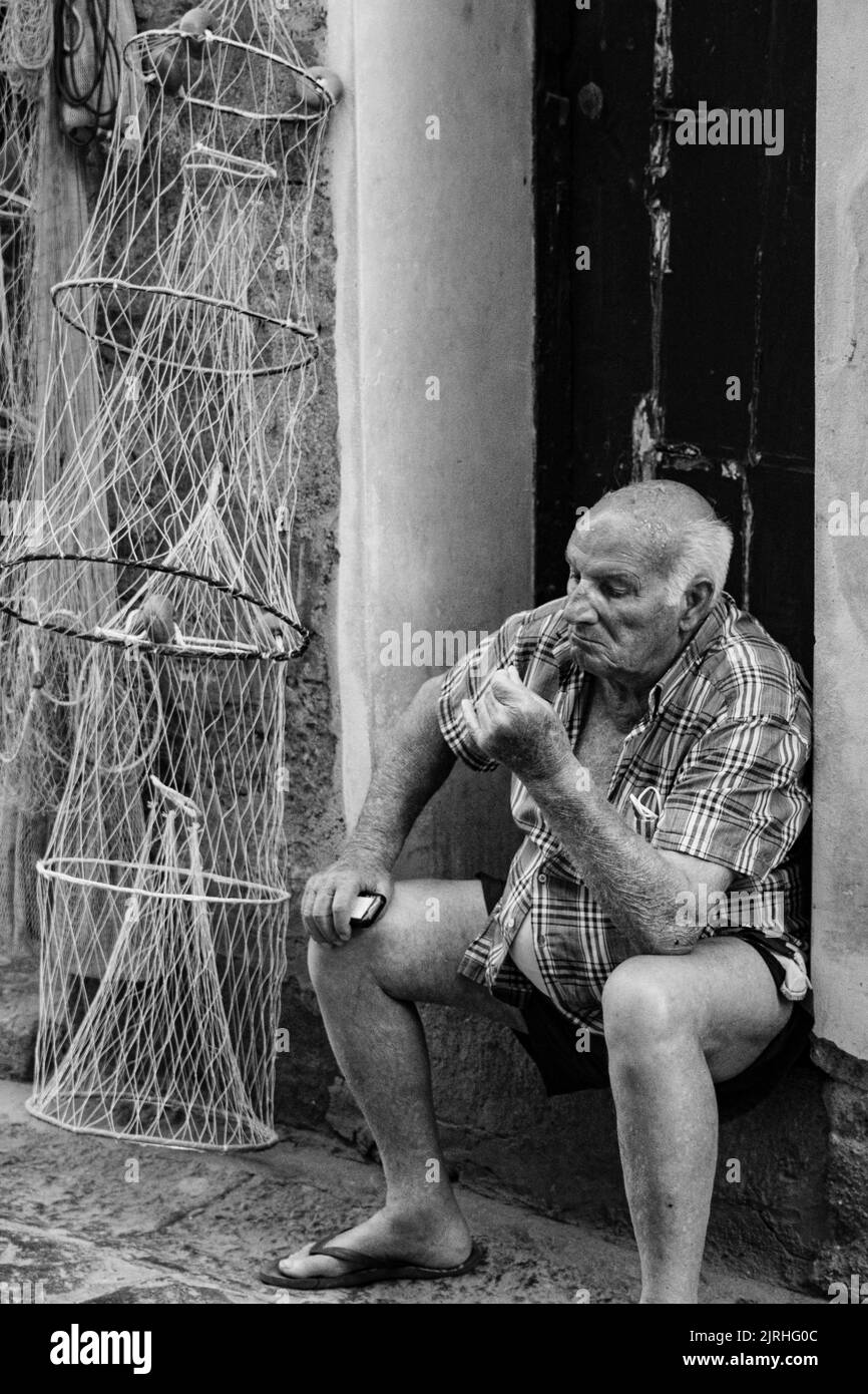 Portrait vertical d'un homme assis à l'entrée de la porte en noir et blanc. Art de rue sicilien: Homme faisant des lumières à partir de filet de pêcheur. Banque D'Images